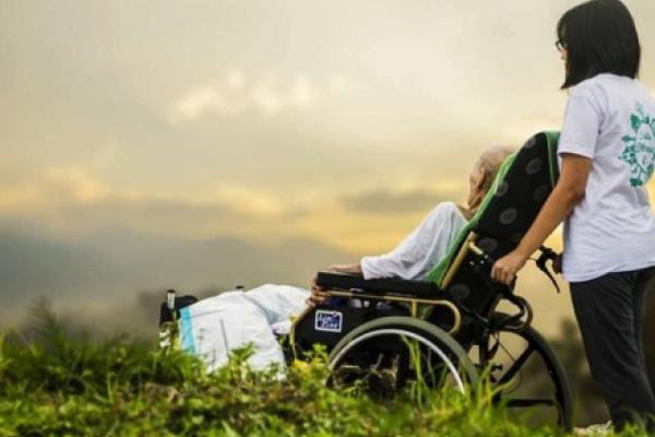 A woman with short dark hair pushes an elderly person in a wheelchair as they look into the distance